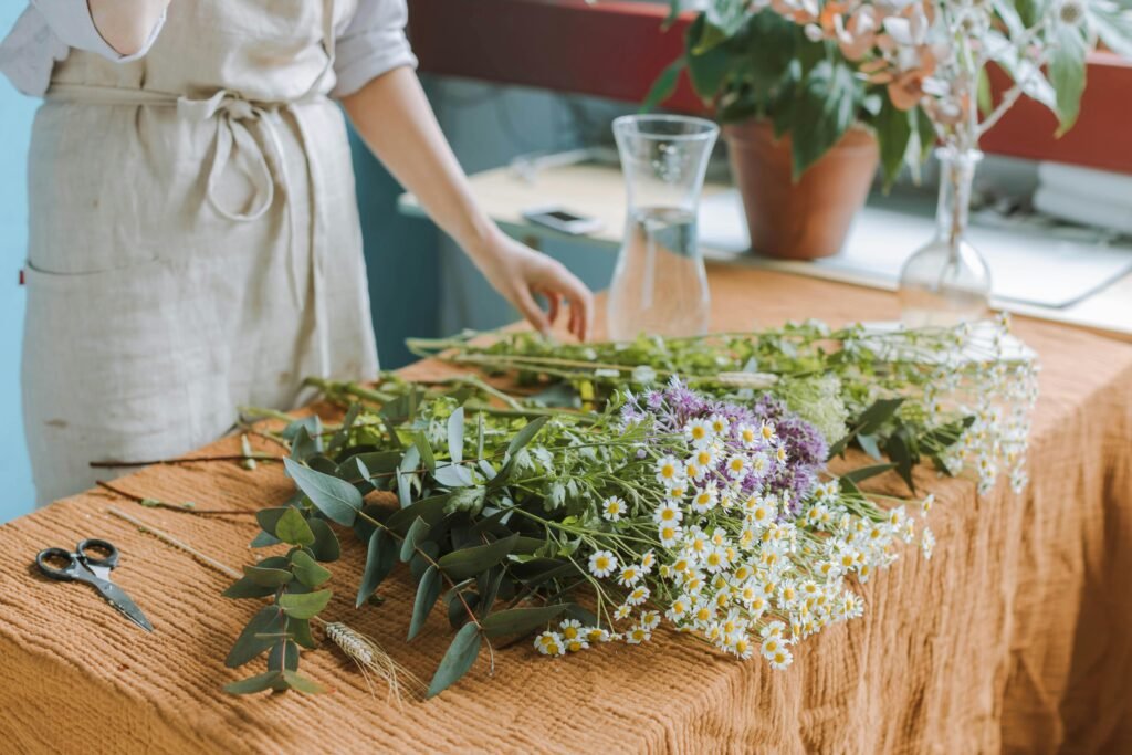 A woman arranges wildflowers on a table indoors with vases and potted plants.